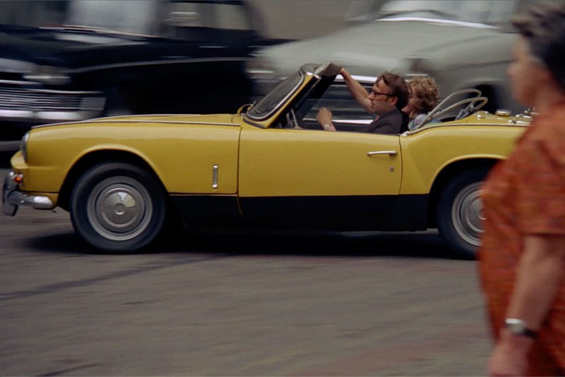 Żółty samochód typu kabriolet z dwiema osobami w środku, jedną za kierownicą, a drugą na siedzeniu pasażera, porusza się ruchliwą ulicą. Na pierwszym planie widać rozmazane samochody i pieszego.