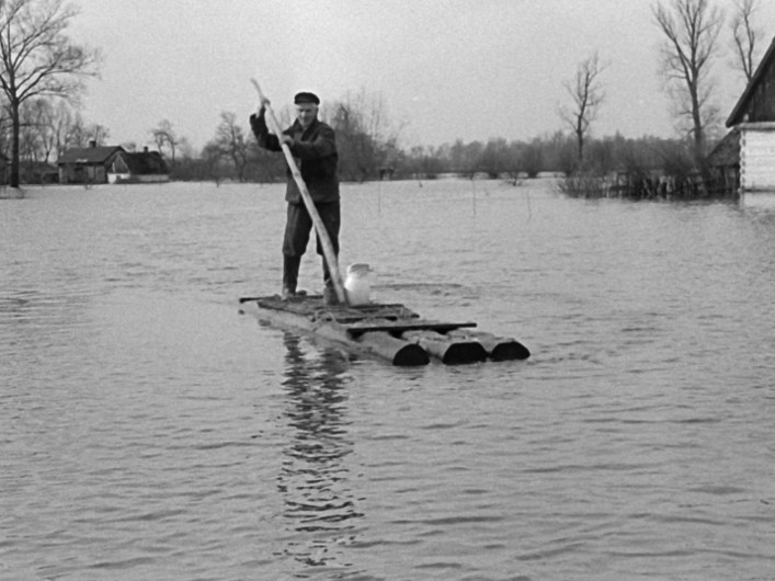 Mężczyzna stoi na prowizorycznej tratwie zrobionej z bali drewnianych i za pomocą długiego kija przemieszcza się przez wodę powodziową. W tle widać częściowo zalane domy i bezlistne drzewa.