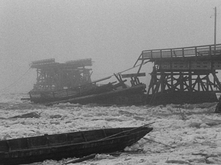 Mglisty widok rzeki z połamanymi drewnianymi odcinkami mostu i kry lodowej; na pierwszym planie widoczna jest uszkodzona łódź.