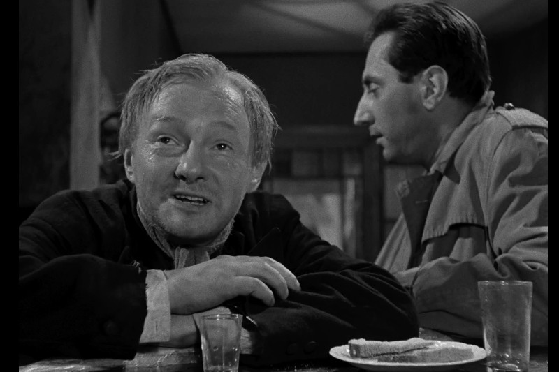 Mężczyzna o jasnych włosach uśmiecha się, opierając się o stół ze szklanką, kieliszkiem do wódki i talerzem z chlebem. Inny mężczyzna w płaszczu siedzi obok niego, odwrócony tyłem. Scena jest czarno-biała.