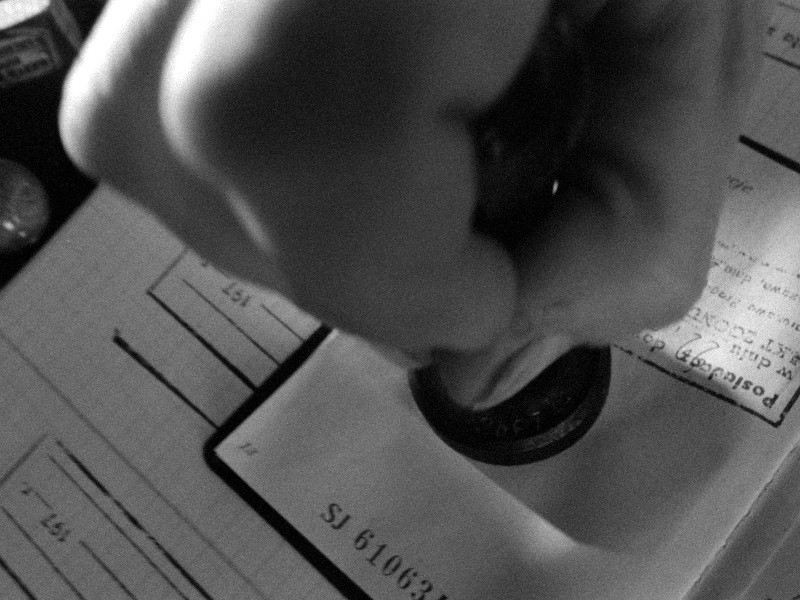 Zbliżenie dłoni stemplującej stronę dokumentu lub paszportu, z widocznymi numerami i tekstem na papierze. Obraz jest czarno-biały.