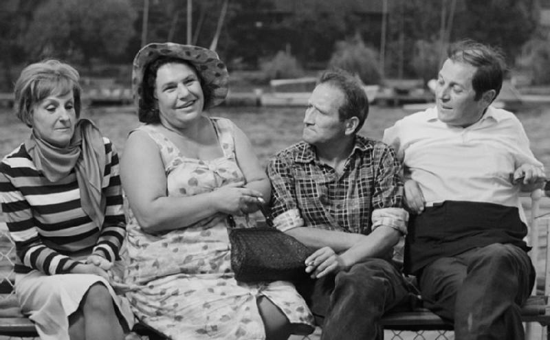 Czterech dorosłych siedzi na ławce na zewnątrz; dwie kobiety po lewej (jedna w pasiastej sukience, druga w kapeluszu i kwiecistej sukience) i dwóch mężczyzn po prawej. Wszyscy wydają się rozmawiać i uśmiechać na czarno-białym zdjęciu.