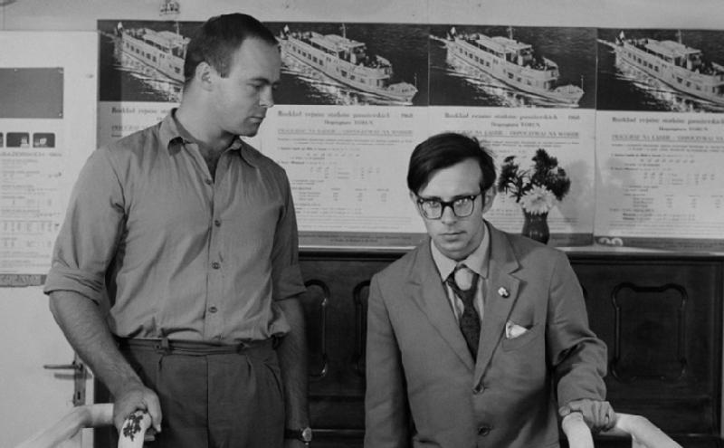 Dwóch mężczyzn stoi w środku; jeden ma na sobie koszulę zapinaną na guziki i trzyma poręcz, a drugi nosi okulary i garnitur. Na ścianie za nimi wiszą plakaty z łodziami i rozkładami jazdy. Scena jest czarno-biała.