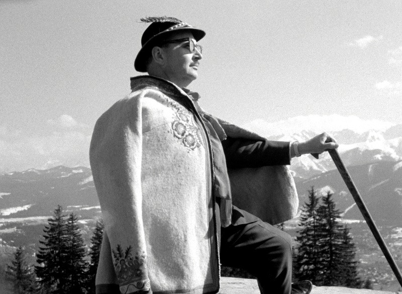 Mężczyzna ubrany w tradycyjny alpejski strój, w tym kapelusz z piórem i haftowany płaszcz, stoi na zewnątrz, trzymając laskę. W tle widać ośnieżone góry i sosny.