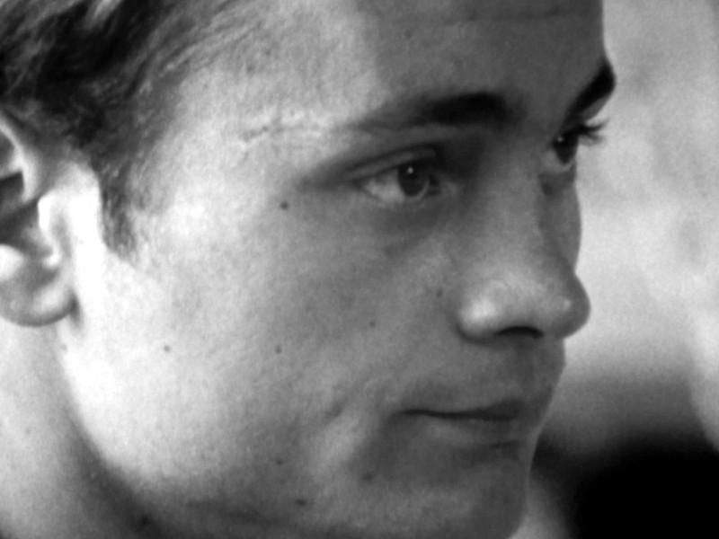 Czarno-białe zbliżenie twarzy młodego mężczyzny, ukazujące poważny lub zamyślony wyraz twarzy. Patrzy lekko w prawo, a delikatne oświetlenie podkreśla jego rysy.