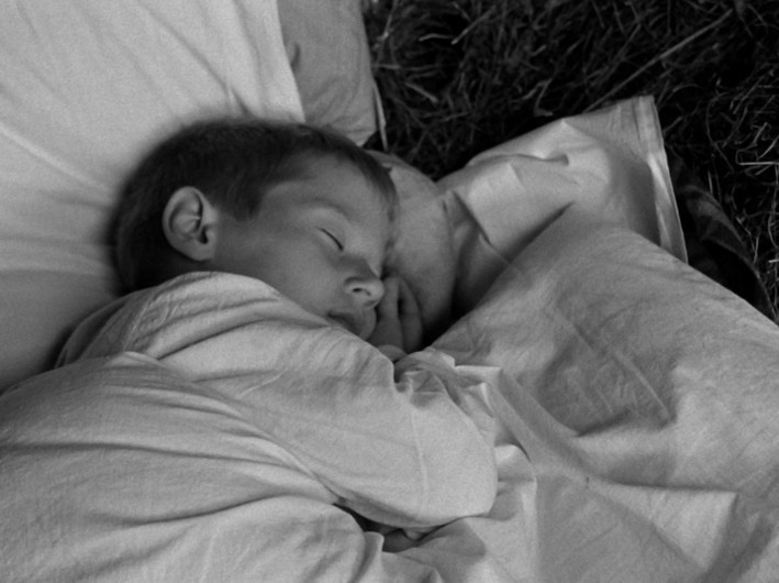 Małe dziecko śpi spokojnie na poduszce, owinięte w koc, z głową opartą na ramieniu. Obraz jest czarno-biały.