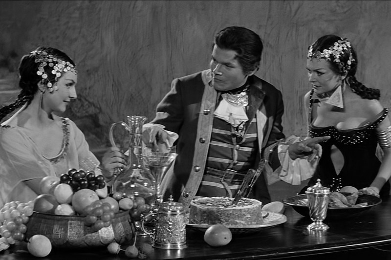 Trzy osoby w historycznych strojach gromadzą się wokół wystawnego stołu z owocami, ciastem, szklankami i srebrnym czajnikiem. Sceneria wydaje się być dramatyczną sceną, z dwiema kobietami i mężczyzną w intensywnej interakcji.