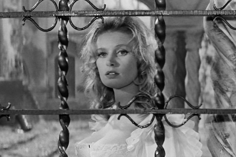 Kobieta o falowanych blond włosach i poważnym wyrazie twarzy stoi za ozdobnymi, poskręcanymi metalowymi prętami, ubrana w jasną sukienkę z odkrytymi ramionami na tle czarno-białej scenerii.