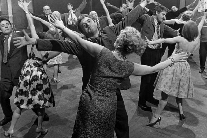Grupa mężczyzn i kobiet w ubraniach w stylu lat 60. tańczy energicznie z wyciągniętymi ramionami na drewnianej podłodze. Scena jest żywa, a tancerze wydają się radośni i ekspresywni.