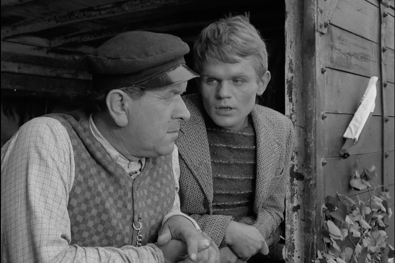 Dwóch mężczyzn w ubraniach vintage wychyla się z drewnianej konstrukcji, rozmawiając. Starszy mężczyzna nosi czapkę i kamizelkę, podczas gdy młodszy mężczyzna w pasiastym swetrze i kurtce patrzy na niego poważnie. Zdjęcie jest czarno-białe.