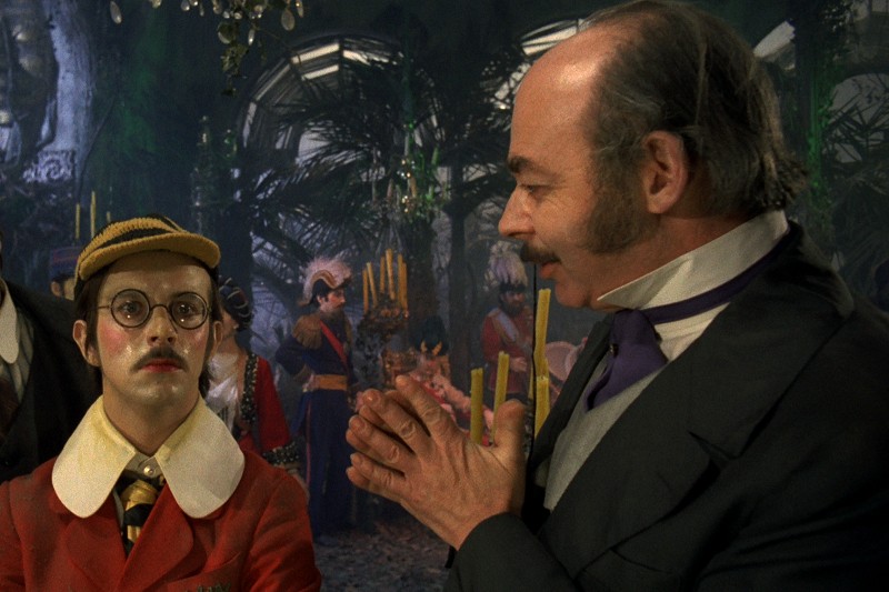 Osoba ubrana w czerwoną kurtkę, żółtą czapkę i okrągłe okulary stoi obok mężczyzny w formalnym stroju z bakami, który z entuzjazmem rozmawia na eleganckim spotkaniu, w tle widać ludzi i świece.