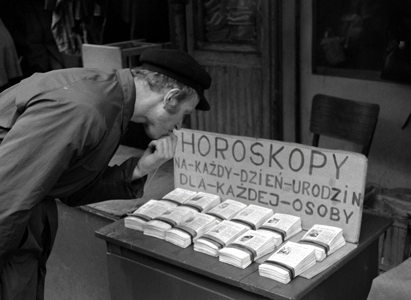 Mężczyzna w płaszczu i kapeluszu pochyla się, by czytać małe otwarte książki wystawione na ulicznym stole. Znak w języku polskim reklamuje horoskopy na każdy dzień urodzin i każdą osobę. W tle widać krzesło i wejście do sklepu.