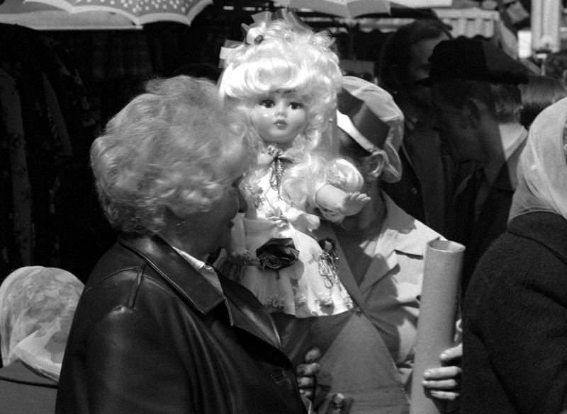 Czarno-białe zdjęcie kobiety trzymającej dużą lalkę z kręconymi, jasnymi włosami, w tłumie ludzi, niektórzy w chustach na głowie i kapeluszach, na targu lub festynie pod gołym niebem.