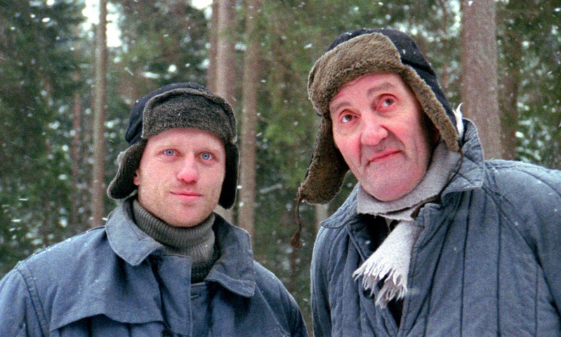 Dwóch mężczyzn ubranych w ciepłe zimowe kurtki i futrzane czapki stoi na zewnątrz w zaśnieżonym lesie, patrząc w kamerę. Płatki śniegu spadają, a obaj wydają się być owinięci w ciepłe ubrania chroniące przed zimnem.