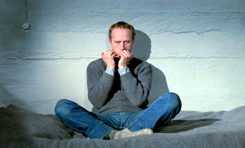 Mężczyzna w szarym swetrze i niebieskich dżinsach siedzi ze skrzyżowanymi nogami na łóżku w skromnym pokoju, grając na harmonijce. Ściana za nim jest szorstka i pusta, a oświetlenie jest słabe.