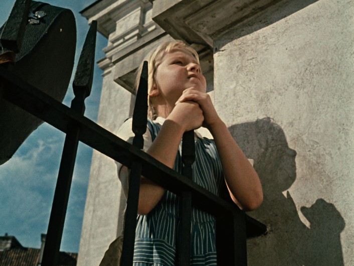 Młoda dziewczyna w pasiastej sukience stoi za czarnym żelaznym płotem, zamyślona spogląda w górę, splecione pod brodą dłonie, rzucając cień na jasną kamienną ścianę.