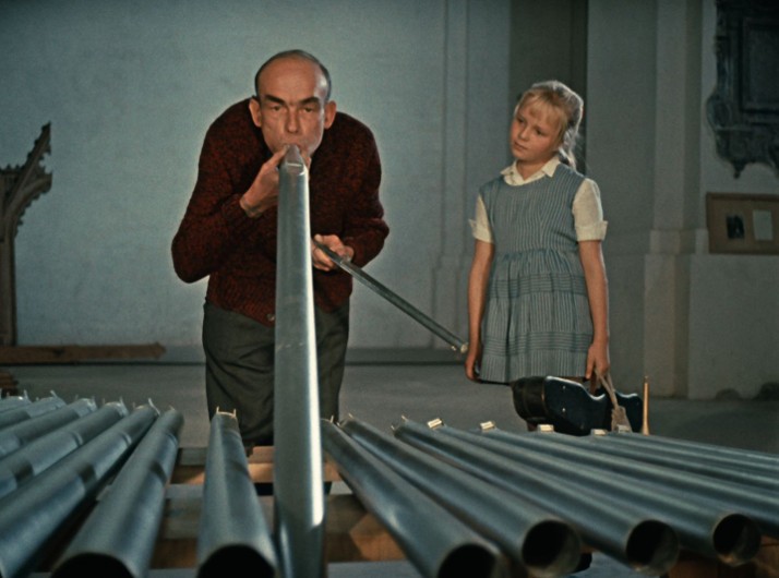 Mężczyzna dmucha w dużą metalową rurę, a młoda dziewczyna w pasiastej sukience obserwuje go, stojąc wśród kilku podobnych rur w pomieszczeniu.