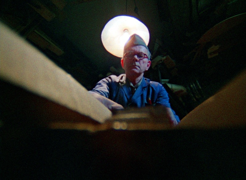 Mężczyzna w niebieskiej koszuli i kapeluszu, ukazany z dołu, oświetlony okrągłą lampą sufitową nad nim, intensywnie pracuje w słabo oświetlonym pomieszczeniu z drewnianymi przedmiotami na pierwszym planie.