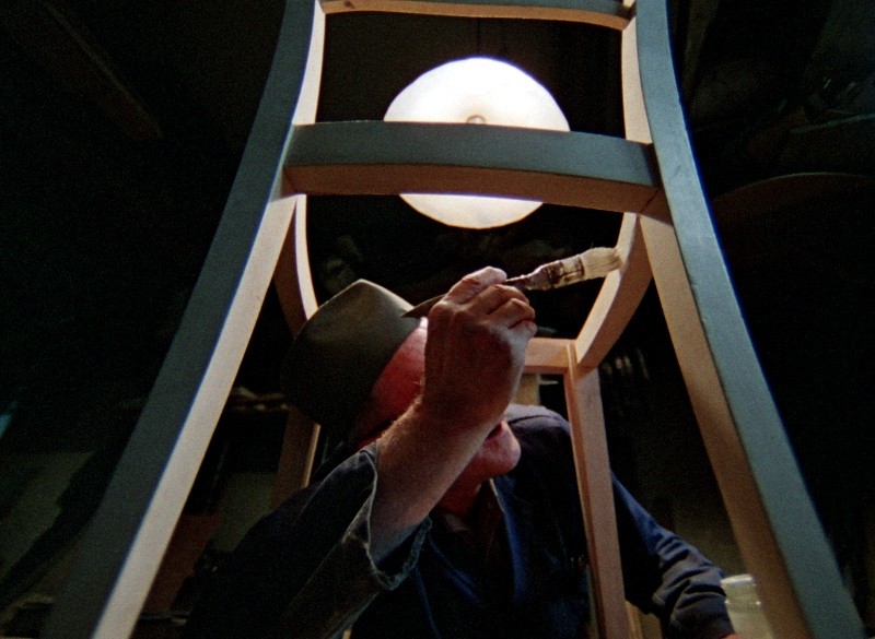 Osoba w kapeluszu maluje pędzlem spodnią część drewnianego krzesła. Widziana jest z niskiego kąta, a nad nią świeci okrągła lampa sufitowa.