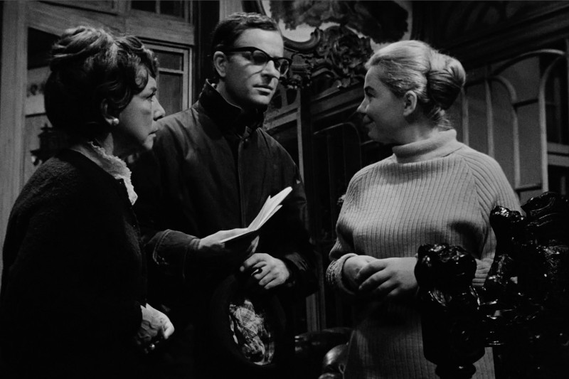 Trzy osoby stoją w pomieszczeniu i rozmawiają; mężczyzna w okularach i ciemnym ubraniu trzyma papiery, naprzeciwko dwóch kobiet, jedna w ciemnym swetrze, a druga w jasnym golfie, w ozdobnym pokoju.