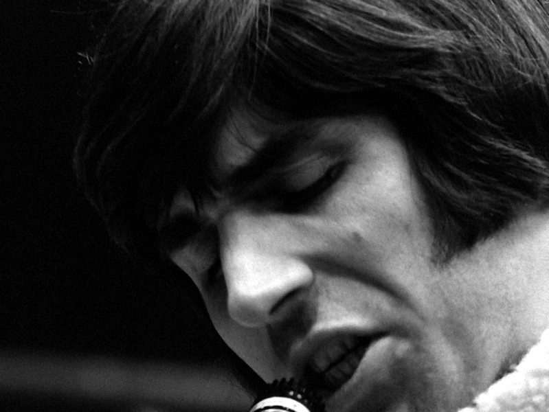 Czarno-białe zdjęcie z bliska przedstawiające osobę z długimi włosami i bakami, śpiewającą lub mówiącą do mikrofonu ze skupionym, intensywnym wyrazem twarzy.