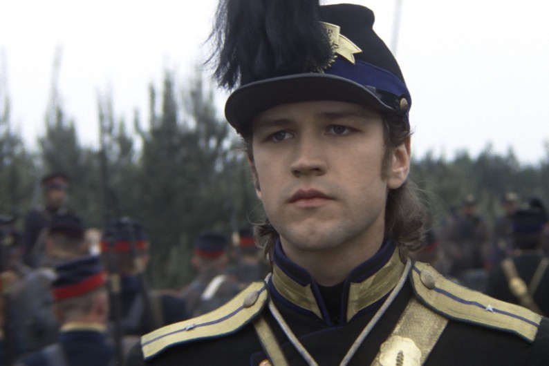 Młody mężczyzna w ciemnym mundurze wojskowym ze złotymi wykończeniami i w czapce z piórami stoi na zewnątrz, a w tle rozmazują się inni żołnierze.