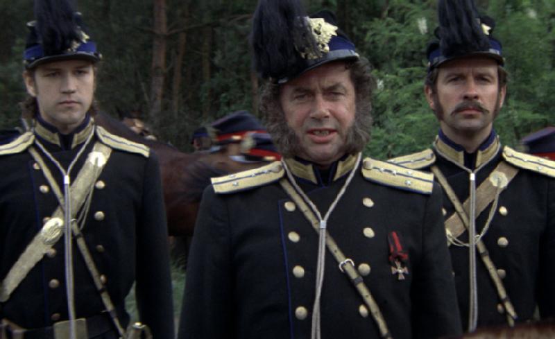 Trzech mężczyzn w ciemnych mundurach wojskowych ze złotymi wykończeniami i kapeluszach z piórami stoi na zewnątrz, patrząc przed siebie. W tle widać drzewa i rozmazane sylwetki. Centralna postać ma charakterystyczne baki.