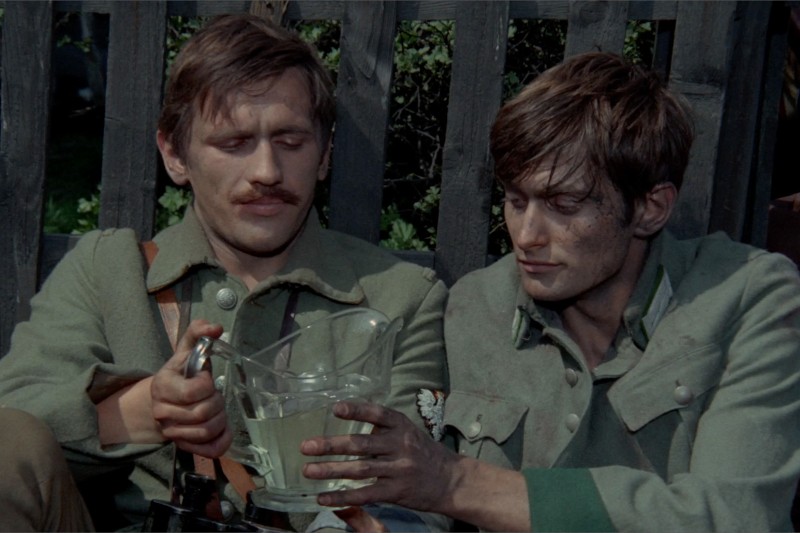 Dwóch mężczyzn w znoszonych zielonych mundurach wojskowych siedzi przy drewnianym płocie. Jeden z nich nalewa wodę z dzbanka do manierki trzymanej przez drugiego, obaj wyglądają na zmęczonych i poważnych.