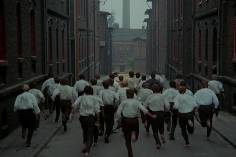 Grupa bosych młodych mężczyzn ubranych w białe koszule i ciemne spodnie biegnie wąską, pustą uliczką między wysokimi ceglanymi budynkami w dzielnicy przemysłowej.