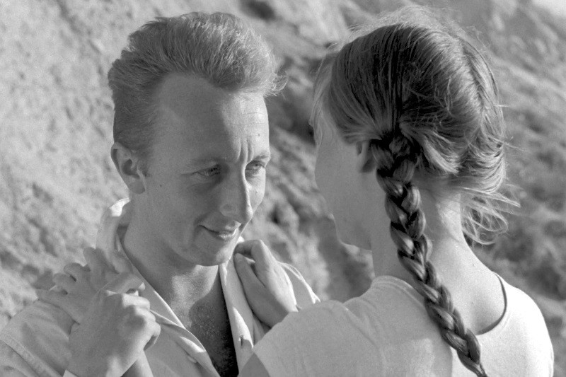 Mężczyzna i kobieta stoją naprzeciw siebie na zewnątrz. Mężczyzna uśmiecha się i trzyma kołnierzyk koszuli, podczas gdy kobieta, widziana od tyłu, ma długi warkocz i lekko trzyma się za ramiona. Tło wydaje się być skalistą powierzchnią.