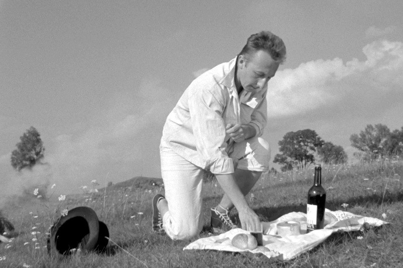 Mężczyzna w jasnym ubraniu klęczy na trawie, przygotowując piknik z chlebem, serem i butelką wina na szmatce. W pobliżu leży kapelusz, a w tle widać drzewa i zachmurzone niebo.