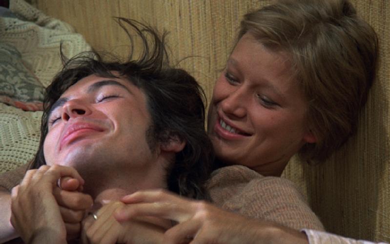 Mężczyzna i kobieta, oboje uśmiechnięci, leżą blisko siebie na kanapie. Kobieta patrzy na mężczyznę czule i żartobliwie trzyma go za ręce, podczas gdy on odchyla się do tyłu i uśmiecha się z zamkniętymi oczami.