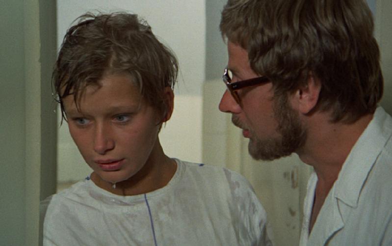 Młoda kobieta z mokrymi włosami i zaniepokojonym wyrazem twarzy stoi obok mężczyzny z brodą i okularami, który wydaje się mówić do niej poważnie. Oboje są w środku, ubrani w jasne ubrania.