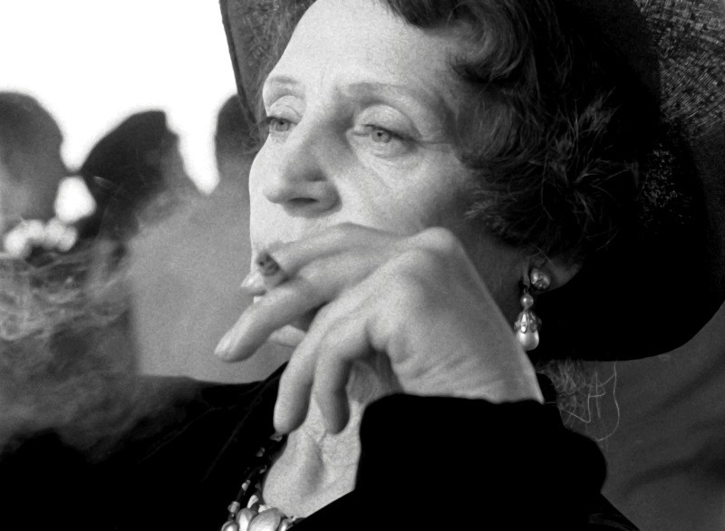 Kobieta w kapeluszu i kolczykach patrzy zamyślona, trzymając papierosa, a dym widoczny jest w pobliżu jej dłoni. Obraz jest czarno-biały, a ona wydaje się zamyślona na rozmytym tle.