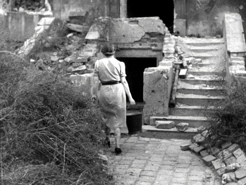 Kobieta w sukience idzie szorstką kamienną ścieżką w kierunku zniszczonego budynku z rozpadającymi się schodami i gruzem, trzymając wiadro w jednej ręce. Scena wydaje się opuszczona i zarośnięta.