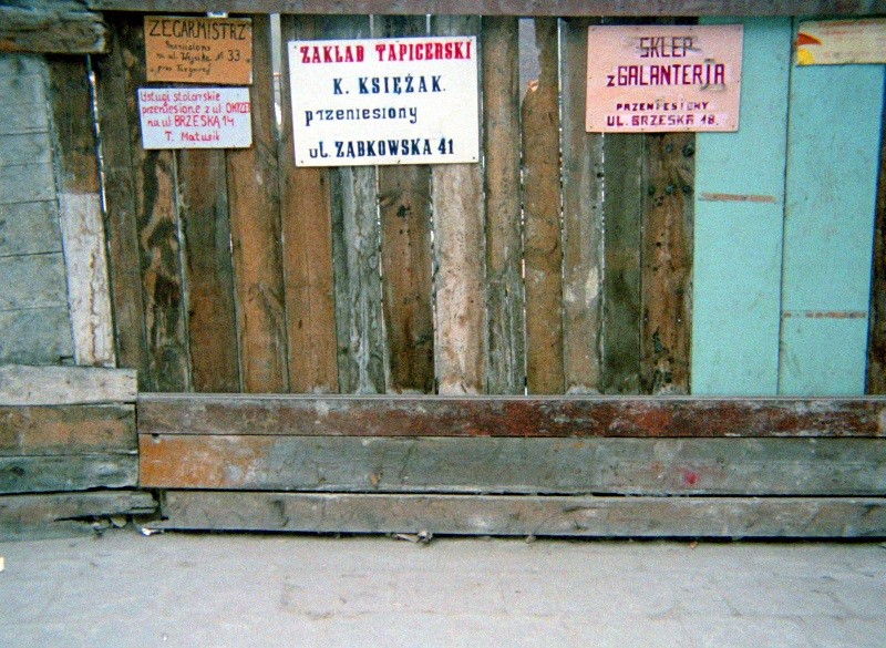 Zwietrzała drewniana ściana z trzema kolorowymi polskimi szyldami reklamującymi różne sklepy i usługi, w tym tapicerkę i pasmanterię, przymocowanymi do nierównych desek. Chodnik poniżej jest popękany i zakurzony.