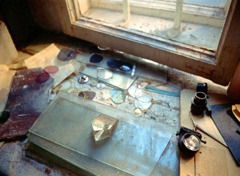 Stary szklany pryzmat, różne soczewki i narzędzia optyczne rozrzucone na zakurzonym parapecie, oświetlone naturalnym światłem z częściowo otwartego okna z kratami. Papiery i tafle szkła dodają scenie naukowego klimatu.