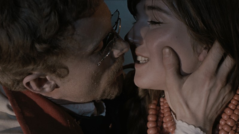 Mężczyzna delikatnie trzyma twarz kobiety, pochylając się blisko, jakby miał ją pocałować. Kobieta uśmiecha się delikatnie, mając na sobie czerwony naszyjnik z koralików. Scena jest intymna, a ciepłe oświetlenie podkreśla ich mimikę.