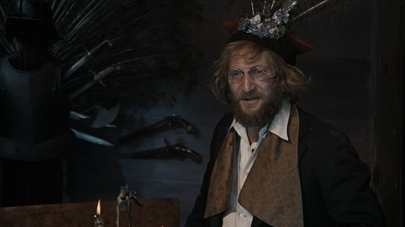 Mężczyzna z brodą i okularami, ubrany w ciemną kurtkę, brązowy szalik i fantazyjny kapelusz ozdobiony kwiatami i pawimi piórami, stoi w pomieszczeniu obok stołu ze świecami i staromodnymi pistoletami na ścianie za nim.