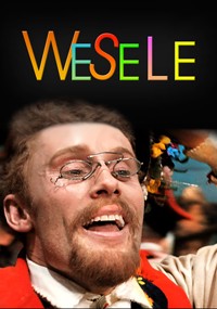 Mężczyzna z wąsami i okularami uśmiecha się radośnie, ubrany w tradycyjny strój, z kolorowymi wstążkami w tle. Słowo WESELE dużymi, wielokolorowymi literami pojawia się nad nim na czarnym tle.