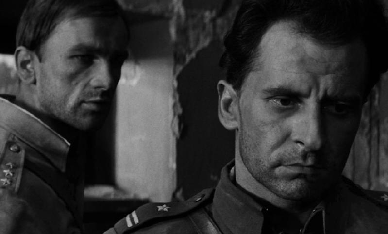 Dwóch mężczyzn w mundurach wojskowych stoi blisko siebie w słabo oświetlonym, zniszczonym pomieszczeniu. Mężczyzna na pierwszym planie patrzy w dół z poważnym wyrazem twarzy, podczas gdy mężczyzna za nim patrzy do przodu, częściowo w cieniu.