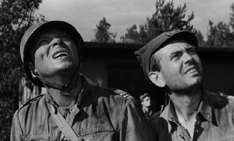 Dwóch mężczyzn w mundurach wojskowych i czapkach patrzy w górę z zaniepokojonymi minami. Drzewa i budynek pojawiają się w tle pod zachmurzonym niebem. Obraz jest czarno-biały.