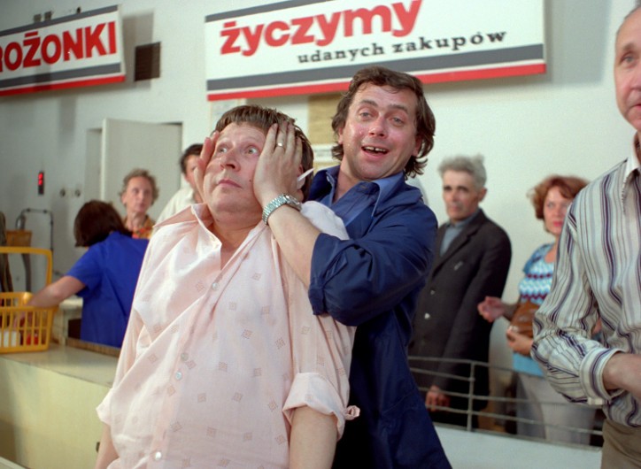 Mężczyzna żartobliwie zakrywa ucho drugiego mężczyzny ręką w kolejce w supermarkecie, podczas gdy ludzie czekają w tle. Scena wydaje się żywa, z widocznymi polskimi znakami i koszykiem na zakupy.