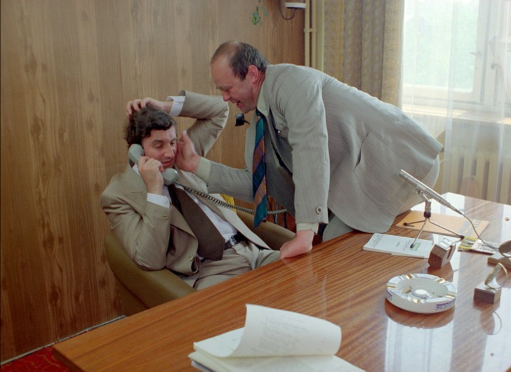 Dwóch mężczyzn w garniturach rozmawia w biurze. Jeden siedzi i rozmawia przez telefon, wyglądając na zaskoczonego, podczas gdy drugi pochyla się, trzymając siedzącego mężczyznę za ucho i uśmiechając się. Na drewnianym biurku leżą dokumenty, telefon i popielniczka.