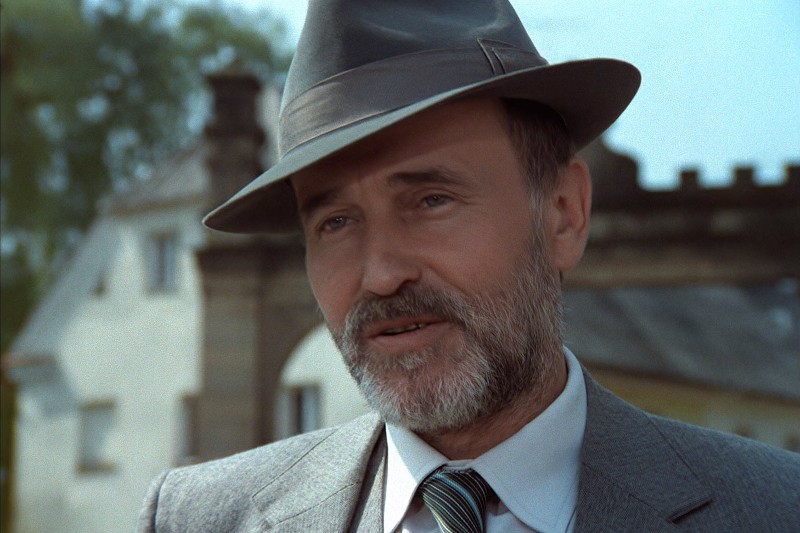 Mężczyzna w średnim wieku z siwą brodą i wąsami, ubrany w szary kapelusz typu fedora i garnitur z krawatem w paski, stoi na zewnątrz przed niewyraźnym kamiennym budynkiem.