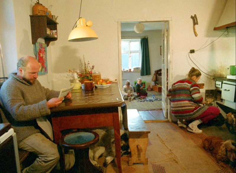 Mężczyzna czyta gazetę przy kuchennym stole, podczas gdy kobieta klęczy przy piecu. W sąsiednim pokoju dzieci bawią się na podłodze. Scena jest przytulna i zagracona, z ciepłym światłem i rustykalnym wystrojem.