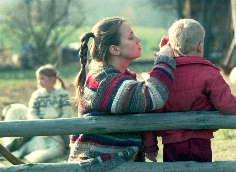 Kobieta w kolorowym swetrze w paski siedzi na drewnianym płocie, delikatnie czyszcząc ucho młodemu blondynowi. Dziewczynka w białym swetrze siedzi rozmazana w tle, a za nimi zielona trawa i drzewa.