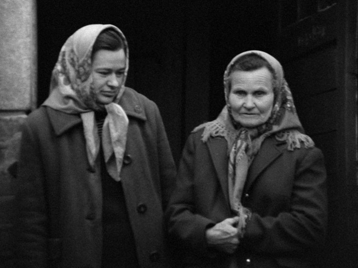 Dwie kobiety w chustach na głowach i płaszczach stoją obok siebie na zewnątrz. Jedna kobieta patrzy lekko w dół, podczas gdy druga patrzy przed siebie, trzymając ręce złożone. Obraz jest czarno-biały.