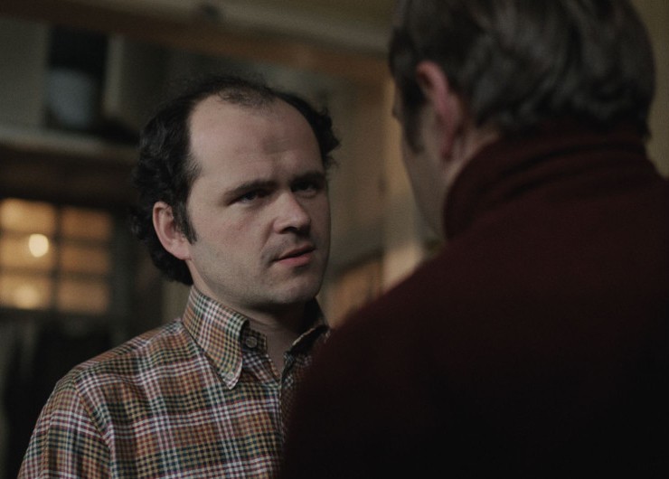Mężczyzna z przerzedzonymi włosami, ubrany w kraciastą koszulę, uważnie przygląda się innej osobie, która stoi tyłem do kamery, w słabo oświetlonym pomieszczeniu.