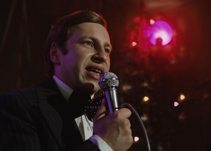 Mężczyzna w garniturze i muszce trzyma mikrofon i przemawia lub śpiewa na scenie. W tle panuje ciepłe oświetlenie, a wokół niego widać różowy reflektor.